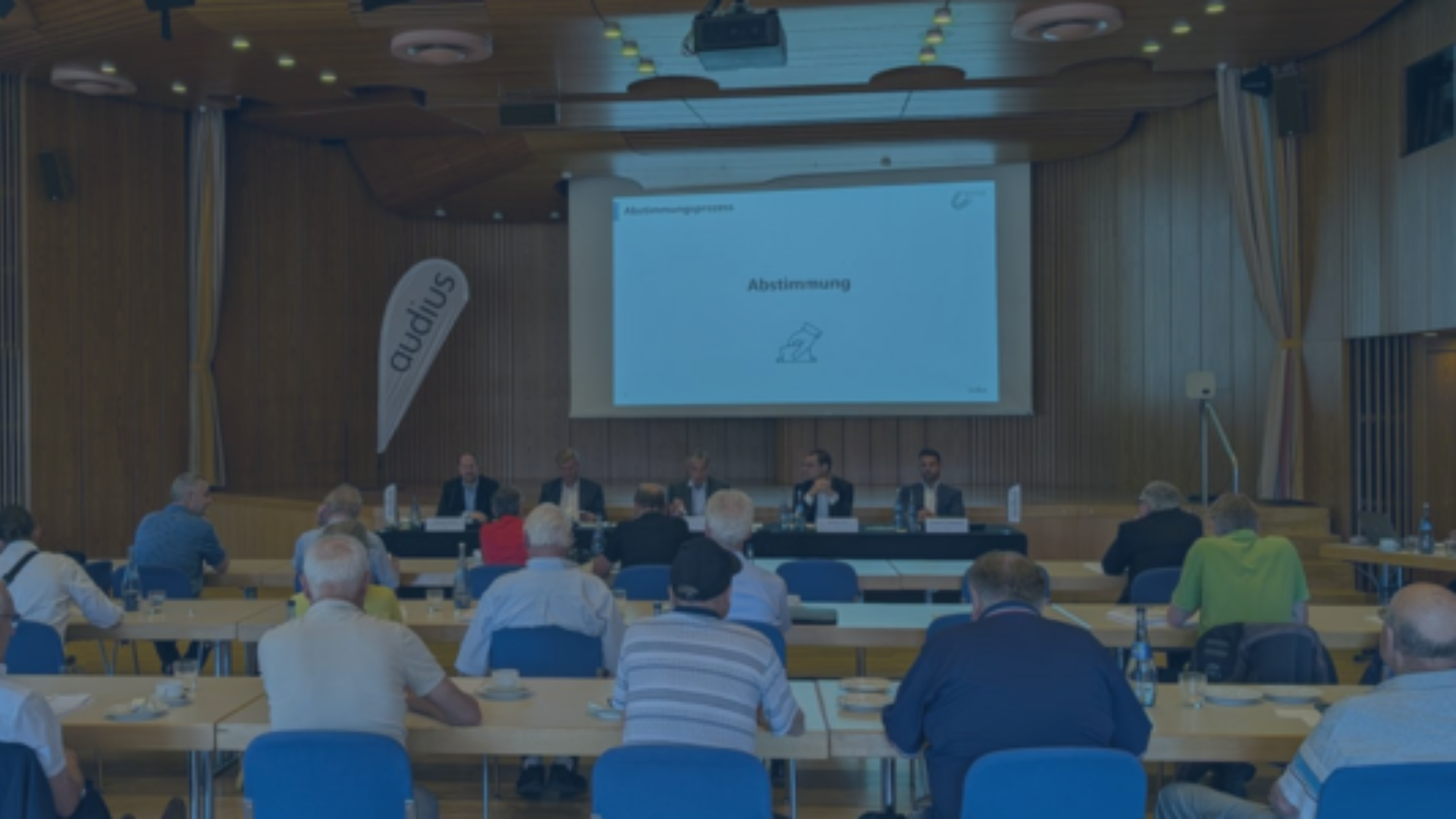 Teilnehmer einer Hauptversammlung sitzen vor einer Bühne mit Podium und Leinwand mit dem Titel „Abstimmung“